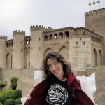 Uma mulher sorridente, magra e pálida, com cabelos castanhos ondulados e óculos, com o capuz do moletom aberto, exibe a camiseta da Sociedade EDS com o símbolo da zebra. A camiseta é preta e o logotipo é branco. O capuz é bordô. Ela está em frente a um castelo com a torre mais alta ostentando três bandeiras: a de Aragão, a espanhola e a europeia. O castelo é cercado por um fosso repleto de grama e arbustos. Trata-se de um palácio medieval fortificado, construído na segunda metade do século XI em estilo mudéjar aragonês, Patrimônio Mundial da UNESCO.